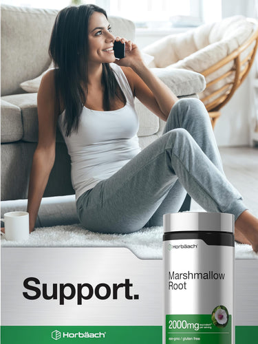 A young adult woman smiles while talking on the phone, sitting on the floor next to a Marshmallow Root bottle.