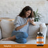 Young woman laughing on a couch, using her phone, with a Chewable Vitamin C bottle beside her.