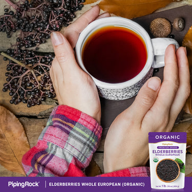 Adult's hands hold a mug of dark drink, surrounded by fresh elderberries and an Elderberries pouch.