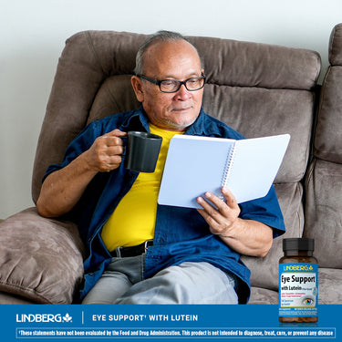 Elderly man sits in a recliner, reading a notebook and holding a mug, next to a bottle of Eye Support with Lutein.