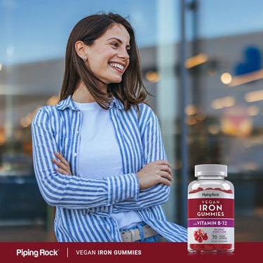 Smiling young woman with arms crossed stands beside a bottle of Vegan Iron Gummies.