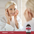 Middle-aged woman smiles and touches her face while looking in a mirror, with a bottle of L-Lysine beside her.