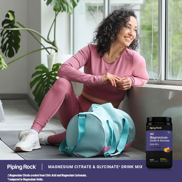 A woman in pink athletic wear sits on a yoga mat by a window, smiling, with a Magnesium Drink Mix.