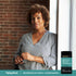 A middle-aged woman smiles by a window with an Ashwagandha + Mag Glycina supplement bottle beside her.