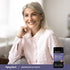 A middle-aged woman smiles while relaxing on a couch with a jar of Magnesium Glycinate powder beside her.