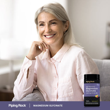 A middle-aged woman smiles while relaxing on a couch with a jar of Magnesium Glycinate powder beside her.