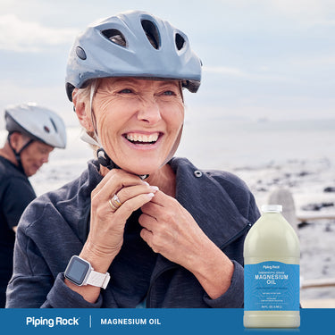 Senior woman smiling and adjusting a bicycle helmet, with a man in the background, next to a Magnesium Oil bottle.