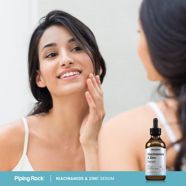 Young woman smiling, touching her cheek in a mirror, next to a Niacinamide & Zinc Serum bottle.