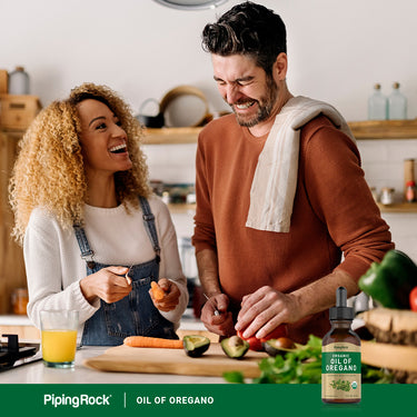 Young couple laughing while chopping vegetables in a kitchen featuring Organic Oil of Oregano bottle.