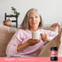 Lifestyle image showing a gray-haired woman relaxing on a couch while holding a white mug, with Piping Rock Mega Multi Women 50+ supplement bottle displayed in the bottom right corner.