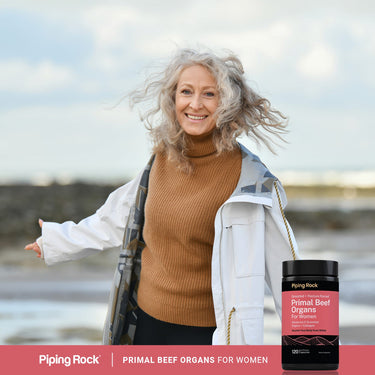 Adult woman smiling outdoors near the water wearing a white jacket and brown sweater, with a bottle of Piping Rock Primal Beef Organs For Women displayed in the lower right corner.