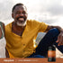 An older man smiling while sitting outdoors next to a Super Ceylon Cinnamon bottle.