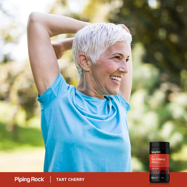 An older woman smiling and stretching outdoors with a bottle of Tart Cherry extract.