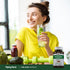 Smiling young woman holding infused water with a Pine Bark Extract bottle on the table in front of her.