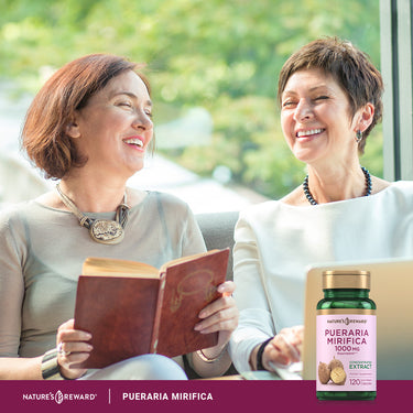 Two middle-aged women laughing and socializing; one holds a book with a Pueraria Mirifica bottle on the table.