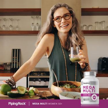A smiling woman over 50 in a kitchen prepares food while holding a green drink next to a Women's Multi 50 Plus bottle.
