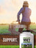 A person in a jacket and hat sits on a wooden fence next to a bottle of Carlyle Grass Fed Whey.
