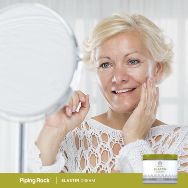 Middle-aged woman smiling at her reflection, touching her cheek with an Elastin Cream jar in view.