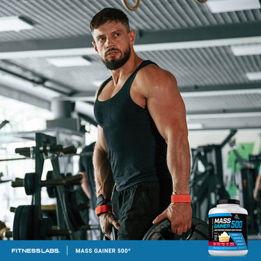 A muscular man exercises in a gym next to a Mass Gainer 500 tub.
