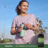 A middle-aged woman smiles while holding a water bottle, next to a bottle of Oil of Oregano with Black Seed Oil.