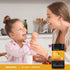 Child smiling while being fed with a spoon by an adult at a kitchen table, with a Piping Rock Kids Multi + Probiotic bottle nearby