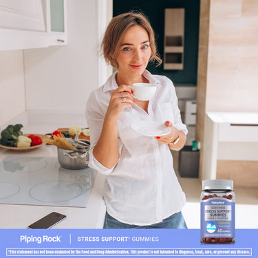 A young woman smiles while holding a teacup in a kitchen next to a bottle of Stress Support Gummies.