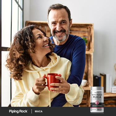 tTwo adults smiling and holding a red mug together indoors with a Piping Rock Red Yellow Black Maca supplement bottle displayed in the bottom right corner