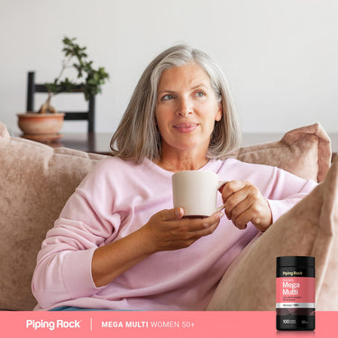 Lifestyle image showing a gray-haired woman relaxing on a couch while holding a white mug, with Piping Rock Mega Multi Women 50+ supplement bottle displayed in the bottom right corner.