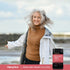 Adult woman smiling outdoors near the water wearing a white jacket and brown sweater, with a bottle of Piping Rock Primal Beef Organs For Women displayed in the lower right corner.