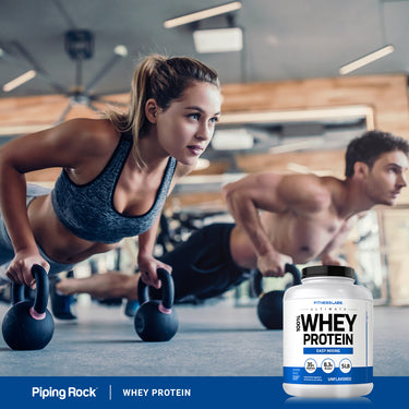 A young woman and man perform push-ups with kettlebells next to a Whey Protein tub, showcasing a fitness lifestyle.